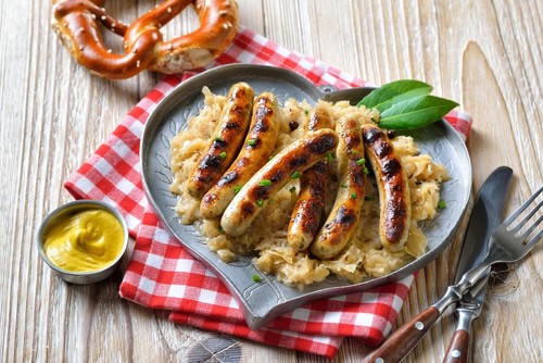 6 Nürnberger Würstchen auf einem herzförmigen Teller mit Sauerkraut arrangiert, unter dem Teller ein rot-weiß-kariertes Tuch und daneben eine Laugenbrezel, Senf und Besteck